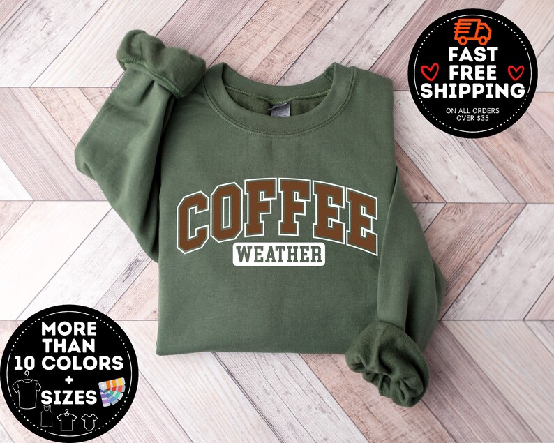 May include: A green sweatshirt with the text "COFFEE WEATHER" in brown letters. The sweatshirt is folded and laid on a wood surface.