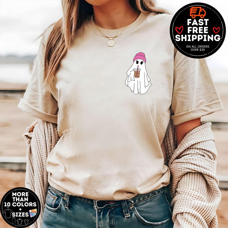 May include: A white ghost wearing a pink beanie hat and holding a cup of iced coffee. The ghost is on a beige t-shirt.