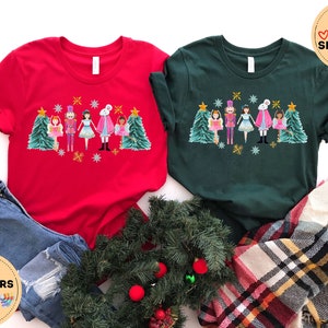 May include: Two red and green t-shirts with a Christmas Nutcracker design. The design features a Nutcracker, a ballerina, a mouse king, and a Christmas tree. The text on the t-shirts says "MORE THAN 10 COLORS + SIZES".