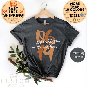 May include: Dark gray heather t-shirt with the text "06 represents freedom 19" in a brown and white design. The shirt is displayed on a wooden hanger. The image also includes text that says "More than 10 colors + sizes".