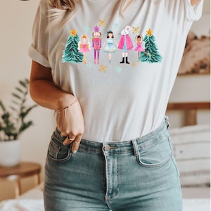 May include: White t-shirt with a watercolor illustration of a nutcracker, a mouse king, and ballerinas in front of Christmas trees. The illustration is in shades of pink, blue, green, and gold.