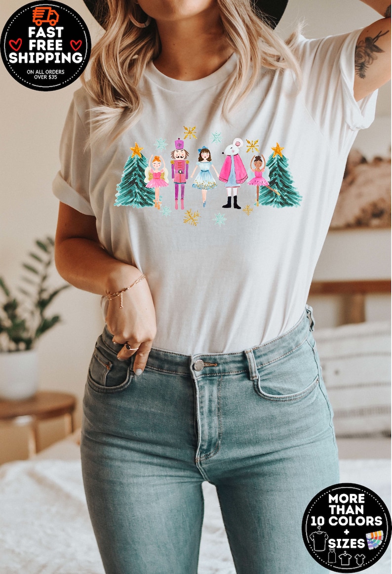 May include: White t-shirt with a watercolor illustration of a nutcracker, a mouse king, and ballerinas in front of Christmas trees. The illustration is in shades of pink, blue, green, and gold.