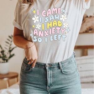 May include: White t-shirt with the text "I CAME I SAW I HAD ANXIETY SO I LEFT" in pastel colors and flower graphics. The shirt is paired with blue jeans. The image also includes a fast, free shipping badge.