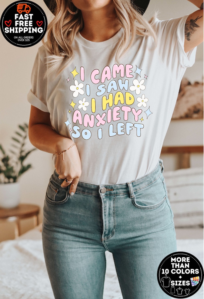 May include: White t-shirt with the text "I CAME I SAW I HAD ANXIETY SO I LEFT" in pastel colors and flower graphics. The shirt is paired with blue jeans. The image also includes a fast, free shipping badge.