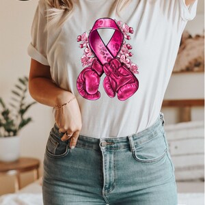 May include: White t-shirt with a pink ribbon and boxing gloves design. The ribbon has a leopard print pattern and is surrounded by pink flowers.  "More than 10 colors + sizes" is written on a circle in the bottom right corner.
