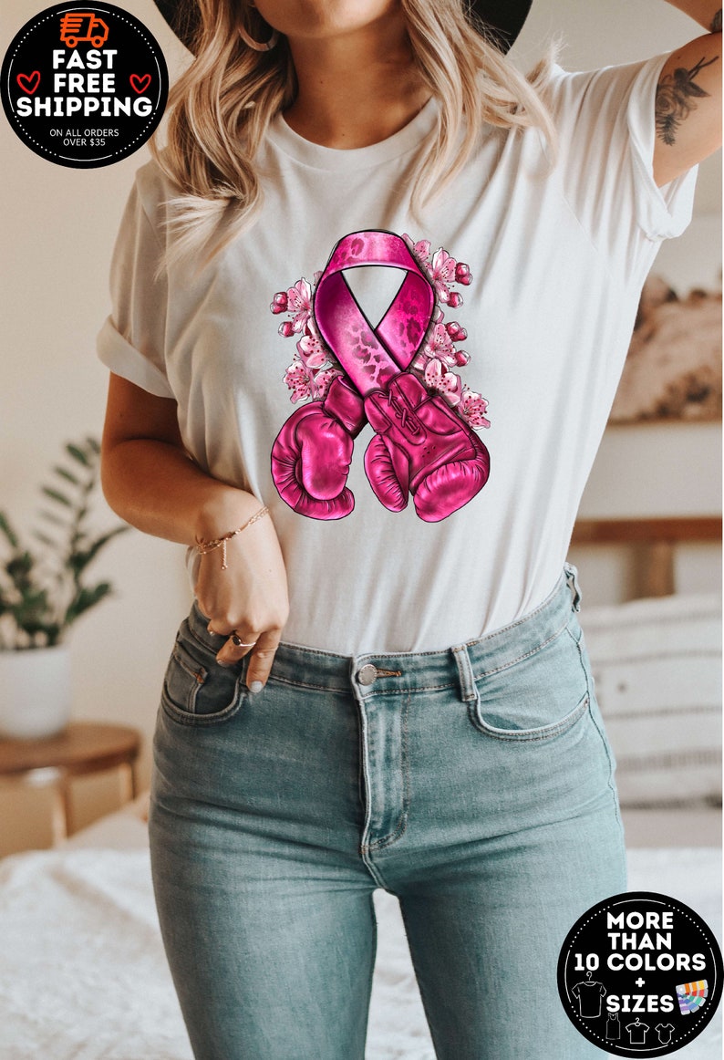 May include: White t-shirt with a pink ribbon and boxing gloves design. The ribbon has a leopard print pattern and is surrounded by pink flowers.  "More than 10 colors + sizes" is written on a circle in the bottom right corner.