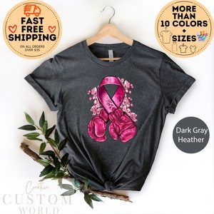 May include: Dark gray heather t-shirt with a pink ribbon and boxing gloves design. The ribbon is surrounded by pink flowers. The text "Creative Custom World" is at the bottom of the image.