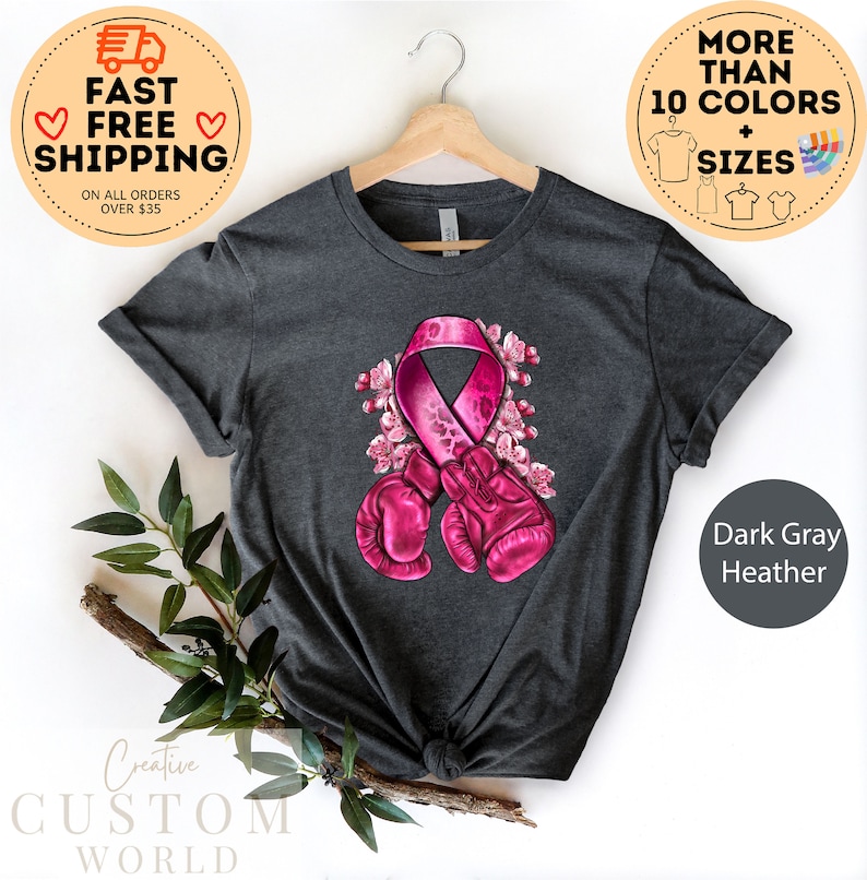 May include: Dark gray heather t-shirt with a pink ribbon and boxing gloves design. The ribbon is surrounded by pink flowers. The text "Creative Custom World" is at the bottom of the image.