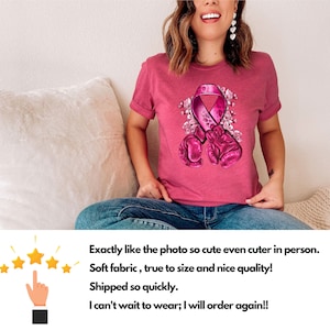May include: Pink t-shirt with a pink ribbon and boxing gloves graphic. The ribbon is surrounded by flowers. The shirt is on a person sitting on a white couch.