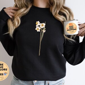 May include: Black sweatshirt featuring a floral design of white and yellow daffodils. The sweatshirt has a crew neck and long sleeves. A white mug with the text "FAST FREE SHIPPING" is held in the hand. The image also includes a graphic with the text "MORE THAN 10 COLORS + SIZES".