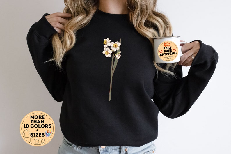 May include: Black sweatshirt featuring a floral design of white and yellow daffodils. The sweatshirt has a crew neck and long sleeves. A white mug with the text "FAST FREE SHIPPING" is held in the hand. The image also includes a graphic with the text "MORE THAN 10 COLORS + SIZES".