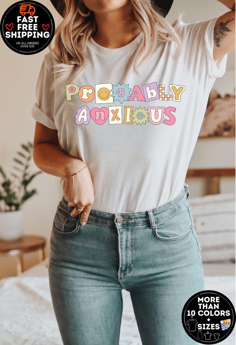 以下が含まれることがあります： カラフルな輪郭のある文字で「PROBABLY ANXIOUS」と書かれた白いTシャツ。各文字は、花、スマイリー、ハートなど、異なるデザインになっています。