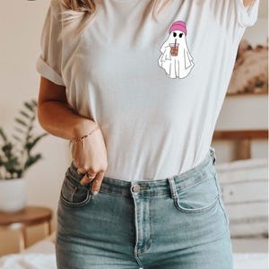 May include: White t-shirt with a graphic of a ghost wearing a pink beanie and holding a cup of iced coffee. The text "More than 10 Colors + Sizes" is visible in the lower right corner of the image.