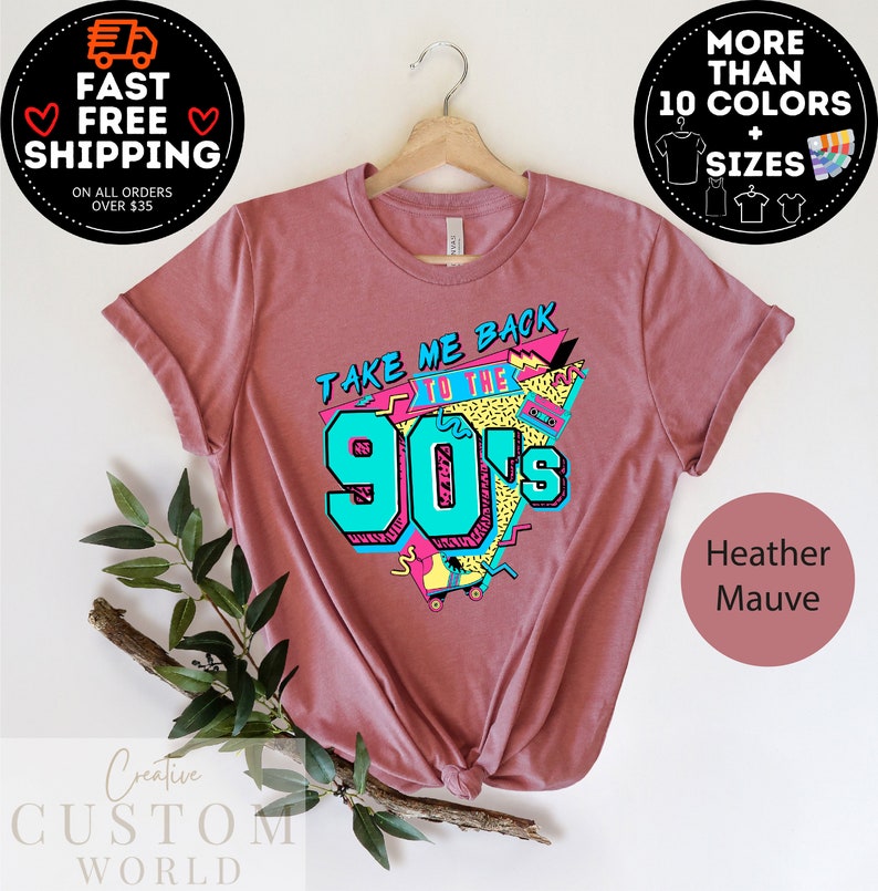 May include: Heather mauve t-shirt with a retro design that says "TAKE ME BACK TO THE 90's" in teal and yellow. The shirt is hanging on a wooden hanger. The image also includes text that says "MORE THAN 10 COLORS + SIZES".