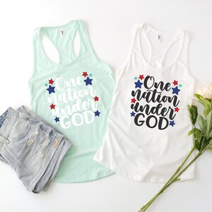 Op de afbeelding: Twee tanktops met de tekst "One nation under GOD" gedrukt in wit op een achtergrond van blauwe, rode en witte sterren. De tanktops liggen op een wit oppervlak.