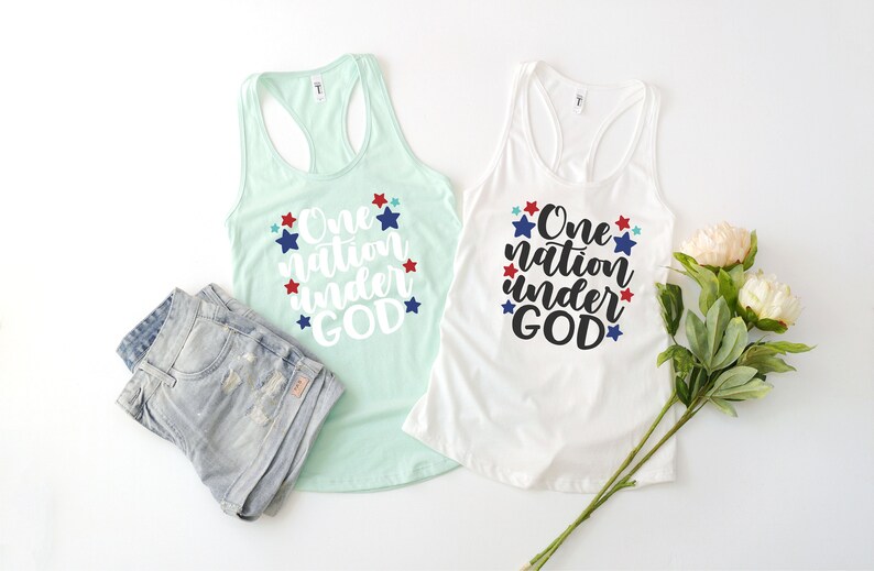 Op de afbeelding: Twee tanktops met de tekst "One nation under GOD" gedrukt in wit op een achtergrond van blauwe, rode en witte sterren. De tanktops liggen op een wit oppervlak.