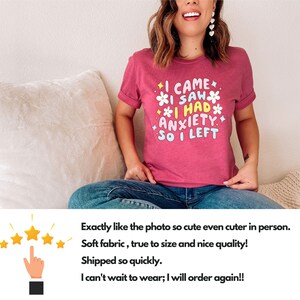 May include: A short-sleeved, magenta t-shirt with the text "I CAME I SAW I HAD ANXIETY SO I LEFT" in a colorful, playful font. The design includes flower and star graphics. The shirt is worn by a person.