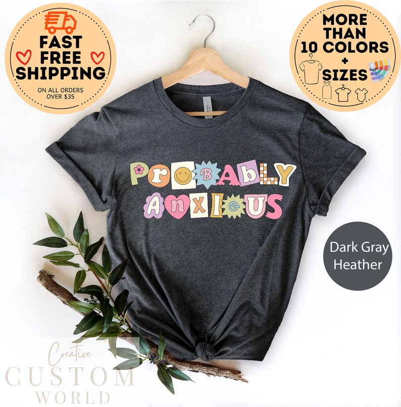以下が含まれることがあります： ダークグレーヘザーのTシャツに、カラフルで遊び心のある文字で「Probably Anxious」と書いてあります。文字は様々なパターンとデザインで飾られています。