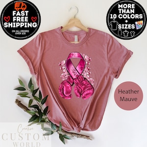 May include: Pink ribbon with a leopard print design and boxing gloves, surrounded by pink flowers. The ribbon is a symbol of breast cancer awareness. The t-shirt is heather mauve.