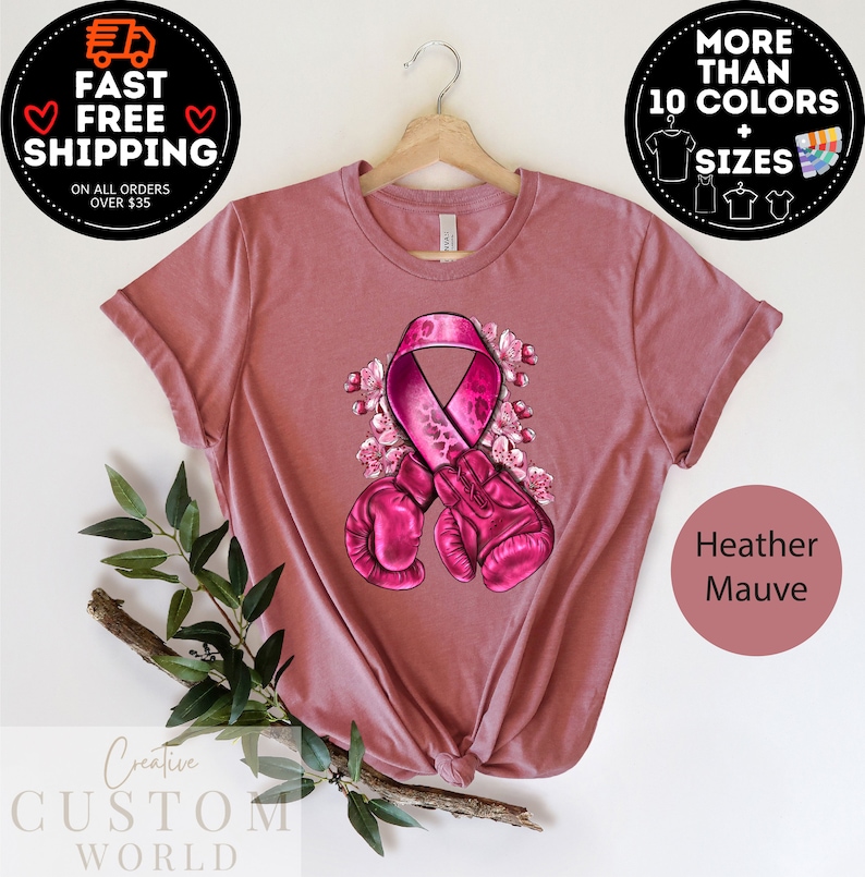 May include: Pink ribbon with a leopard print design and boxing gloves, surrounded by pink flowers. The ribbon is a symbol of breast cancer awareness. The t-shirt is heather mauve.