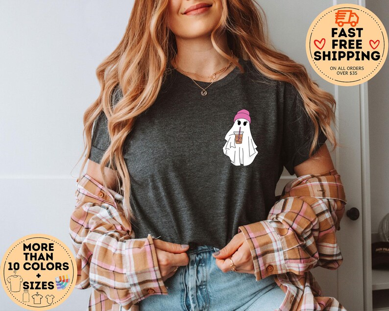 May include: A dark gray t-shirt with a graphic of a ghost wearing a pink beanie and holding a cup of iced coffee. The text "More Than 10 Colors + Sizes" is on a circle sticker in the bottom left corner of the image.
