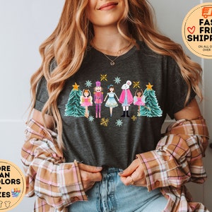 May include: A dark grey t-shirt with a colorful illustration of a nutcracker, a ballerina, and a white rabbit with a Christmas tree on each side. The illustration is surrounded by snowflakes and stars.