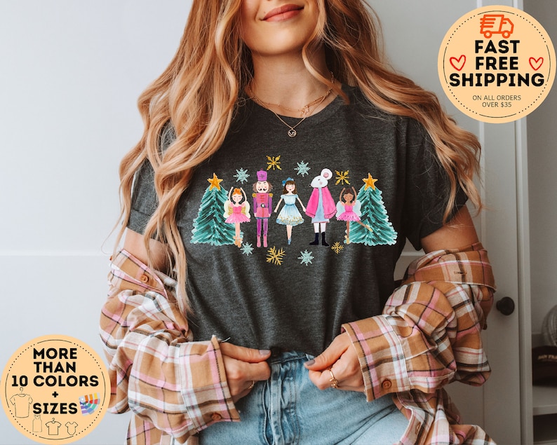 May include: A dark grey t-shirt with a colorful illustration of a nutcracker, a ballerina, and a white rabbit with a Christmas tree on each side. The illustration is surrounded by snowflakes and stars.