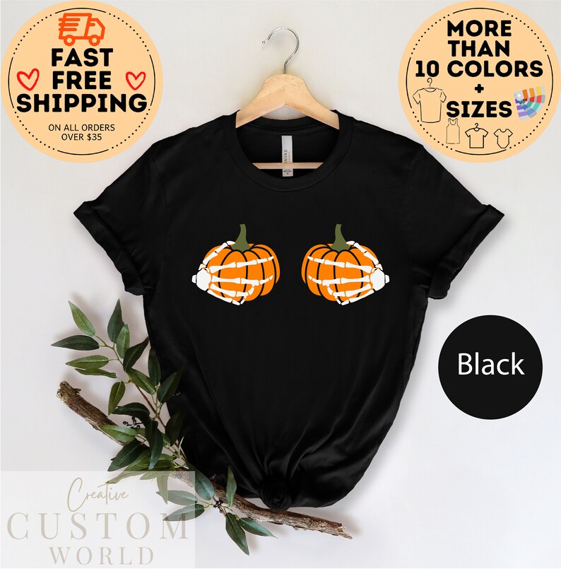 May include: Black t-shirt with a graphic of two orange pumpkins with skeleton hands. The text "More Than 10 Colors + Sizes" is in a circle above the shirt. The text "Black" is in a circle below the shirt.