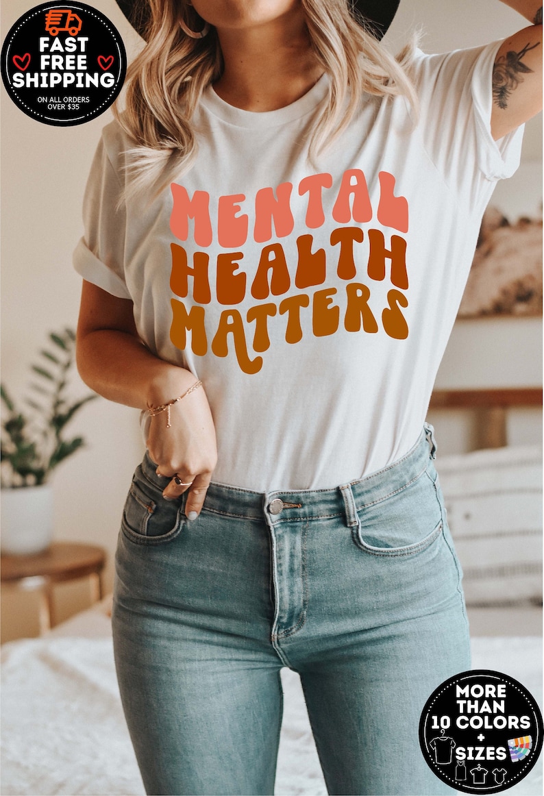 May include: White t-shirt with the text "MENTAL HEALTH MATTERS" in a retro, rainbow-colored font.