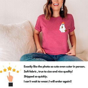 May include: A woman wearing a pink t-shirt with a white ghost graphic holding a cup of coffee and a pumpkin. The ghost is wearing an orange beanie. The text on the t-shirt reads "Exactly like the photo so cute even cuter in person. Soft fabric, true to size and nice quality! Shipped so quickly. I can't wait to wear; I will order again!!"