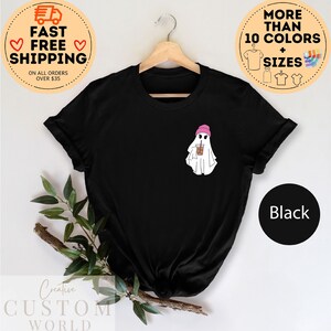 May include: Black t-shirt with a white ghost wearing a pink beanie and holding a brown and white cup. The text "More Than 10 Colors + Sizes" is in a circle above the shirt.