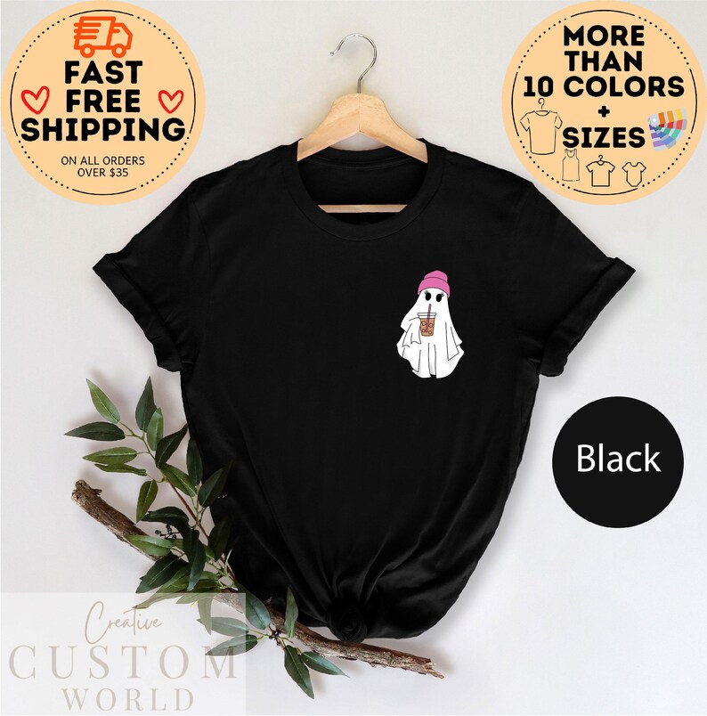 May include: Black t-shirt with a white ghost wearing a pink beanie and holding a brown and white cup. The text "More Than 10 Colors + Sizes" is in a circle above the shirt.