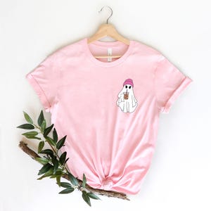 May include: Pink t-shirt with a white ghost wearing a pink beanie and holding a brown iced coffee. The ghost is on the left side of the shirt.