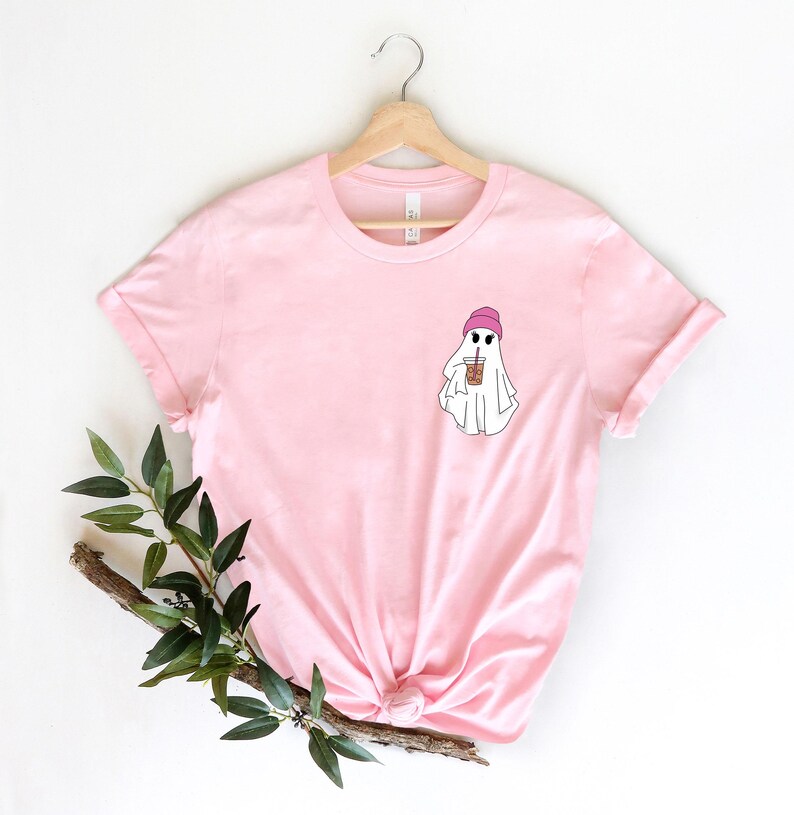 May include: Pink t-shirt with a white ghost wearing a pink beanie and holding a brown iced coffee. The ghost is on the left side of the shirt.