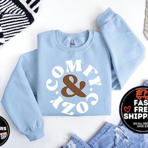 May include: A light blue sweatshirt with a white circle graphic that says "Comfy & Cozy" in a retro font. The ampersand is brown.
