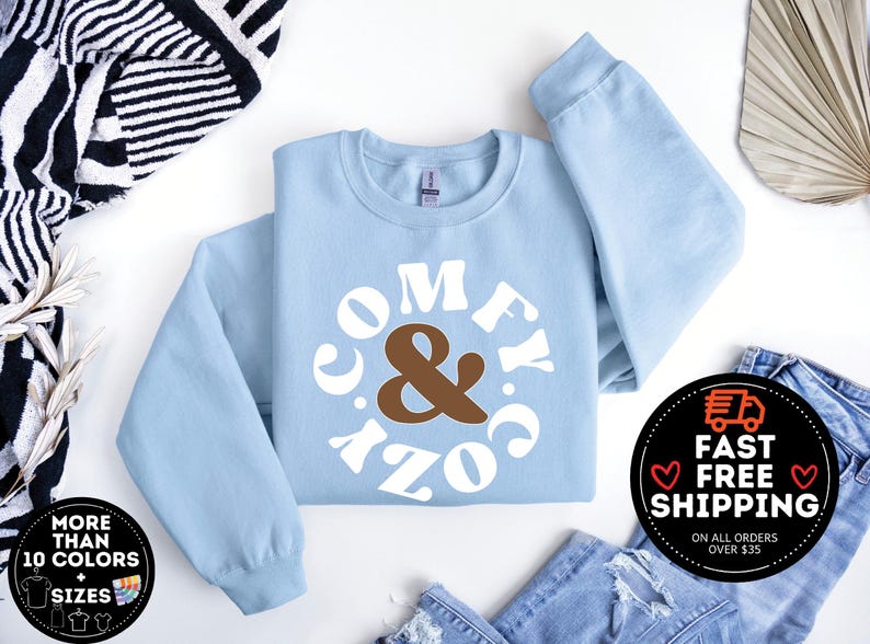 May include: A light blue sweatshirt with a white circle graphic that says "Comfy & Cozy" in a retro font. The ampersand is brown.