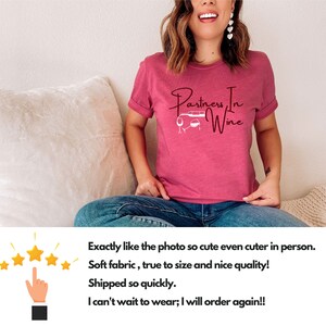 Puede incluir: Una camiseta rosa con el texto "Partners In Wine" en letra cursiva. El texto est&aacute; acompa&ntilde;ado por una peque&ntilde;a ilustraci&oacute;n de un coche con dos copas de vino.