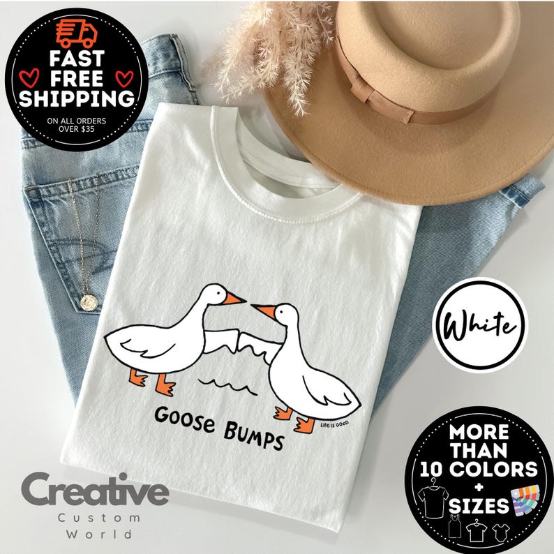 以下が含まれることがあります： 白いTシャツに、2羽のガチョウのグラフィックと「Goose Bumps」と「Life is good」の文字がプリントされています。Tシャツはデニムの背景に茶色の帽子を被せてディスプレイされています。