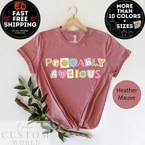 以下が含まれることがあります： ヘザーモーヴのTシャツに、カラフルな文字で「ProBabY ANXIOUS」と書いてあります。文字は花、スマイリーフェイス、星で飾られています。