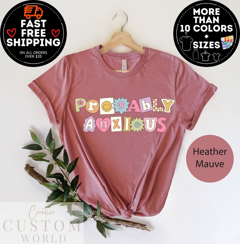 以下が含まれることがあります： ヘザーモーヴのTシャツに、カラフルな文字で「ProBabY ANXIOUS」と書いてあります。文字は花、スマイリーフェイス、星で飾られています。
