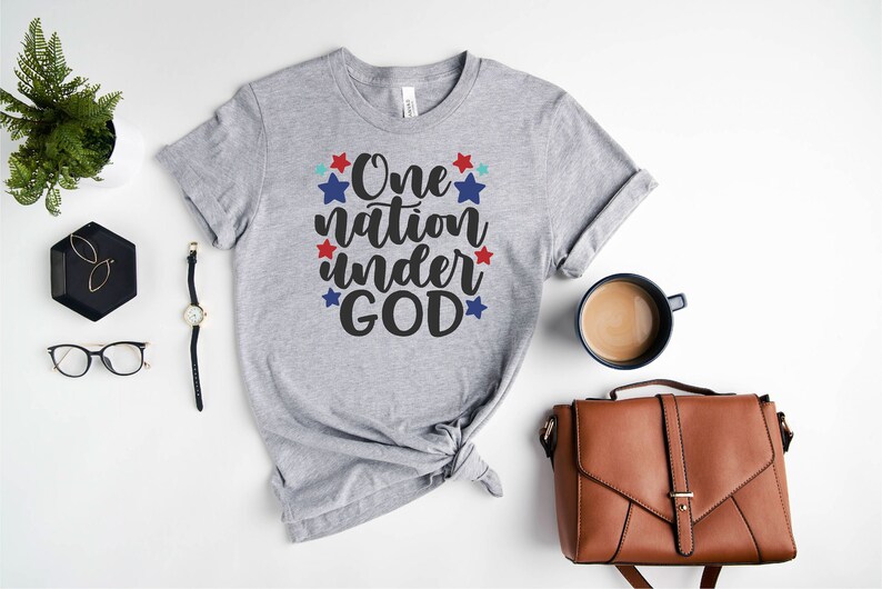 Op de afbeelding: Een grijs T-shirt met de tekst "One nation under GOD" in zwarte letters en rode en blauwe sterren.