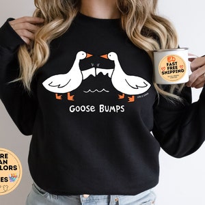 Puede incluir: Sudadera negra con un gr&aacute;fico de dos gansos blancos que se miran con el texto "Goose Bumps". La modelo lleva la sudadera y sostiene una taza blanca con el texto "Env&iacute;o r&aacute;pido y gratuito".