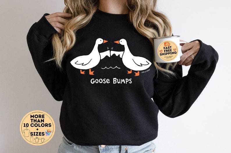 Puede incluir: Sudadera negra con un gr&aacute;fico de dos gansos blancos que se miran con el texto "Goose Bumps". La modelo lleva la sudadera y sostiene una taza blanca con el texto "Env&iacute;o r&aacute;pido y gratuito".