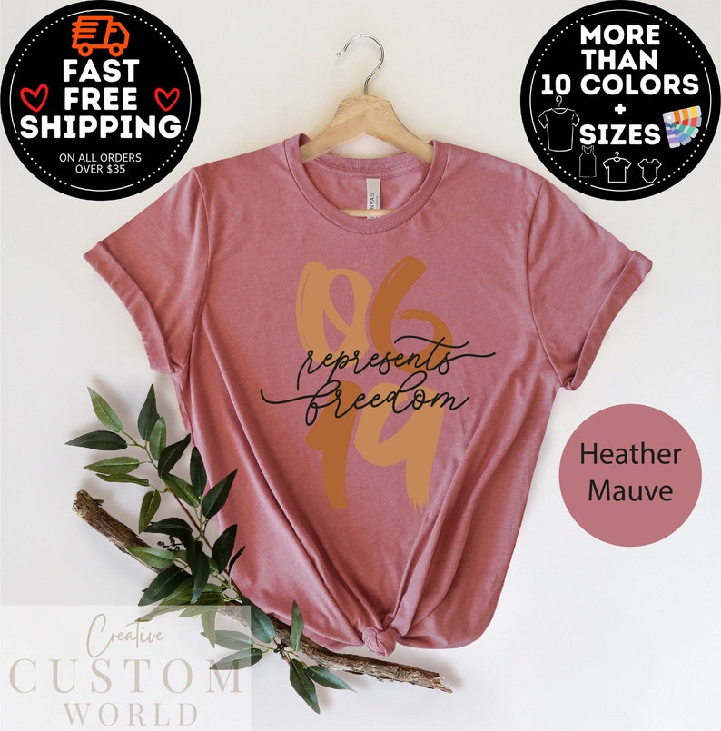 May include: Heather mauve t-shirt with the text "96 represents freedom" in a stylized design. The shirt is displayed on a wooden hanger. Additional text includes "More than 10 colors + sizes" and "Fast Free Shipping".