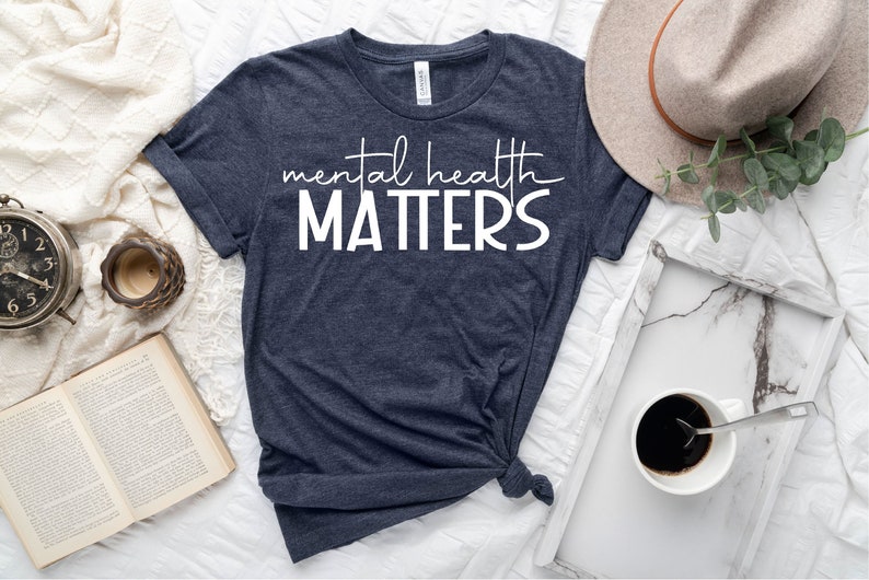 May include: A navy blue t-shirt with white text that reads "mental health MATTERS". The shirt is laid out on a white surface with a book, a clock, a cup, a hat, and a tray with a cup of coffee.