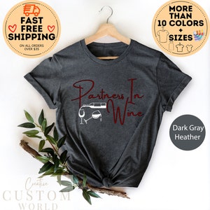 Puede incluir: Camiseta de color gris oscuro jaspeado con el texto "Partners In Wine" en letras rojas. El dise&ntilde;o incluye una silueta blanca de una botella de vino que se vierte en una copa.