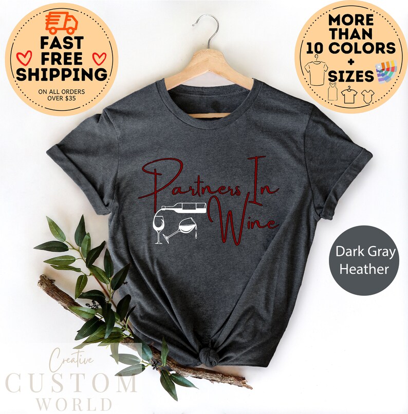 Puede incluir: Camiseta de color gris oscuro jaspeado con el texto "Partners In Wine" en letras rojas. El dise&ntilde;o incluye una silueta blanca de una botella de vino que se vierte en una copa.