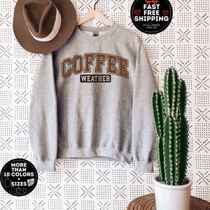 May include: Gray sweatshirt with the text "COFFEE WEATHER" in brown letters. The sweatshirt is hanging on a wooden hanger against a white background with a black and white geometric pattern.