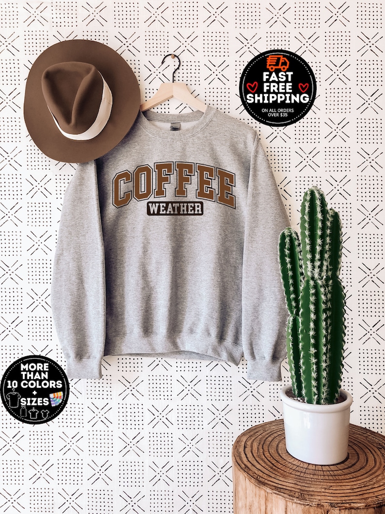 May include: Gray sweatshirt with the text "COFFEE WEATHER" in brown letters. The sweatshirt is hanging on a wooden hanger against a white background with a black and white geometric pattern.