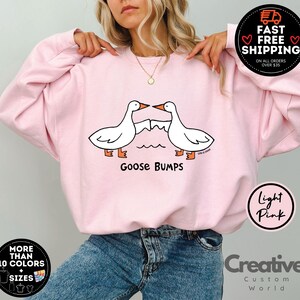 Puede incluir: Sudadera rosa claro con un gr&aacute;fico de dos gansos bes&aacute;ndose y el texto "Goose Bumps".